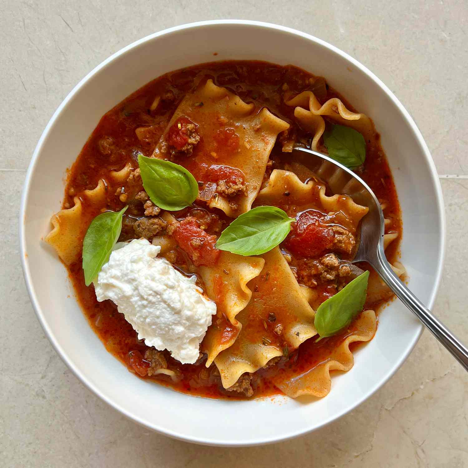 A white bowl filled with a red broth studded with tomato chunks and ground beef, lasagna noodles, and topped with a dollop of ricotta and a few basil leaves