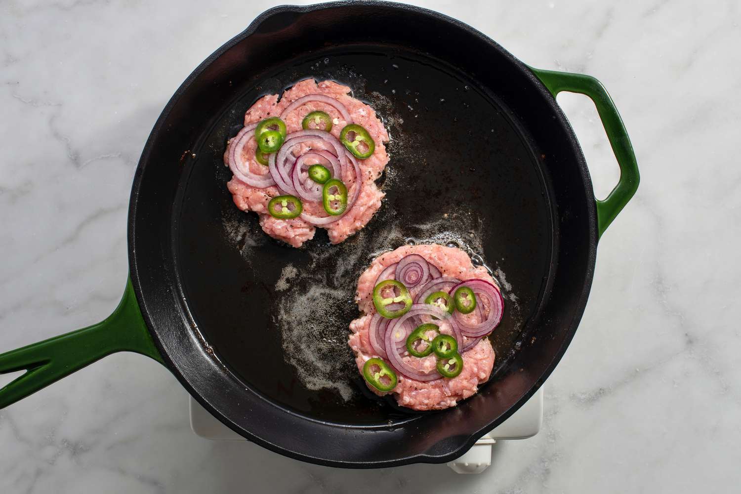 Flattened chicken patties topped with onions and jalapenos cooking in a cast iron skillet