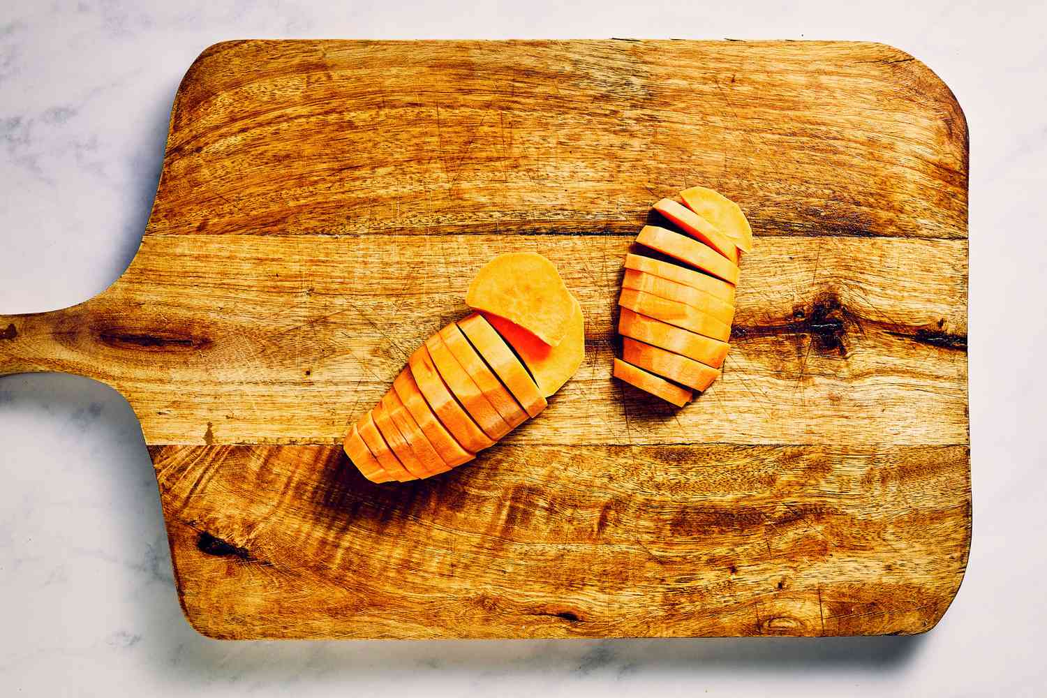 A wood cutting board with a halved, peeled sweet potatoes sliced into half moons