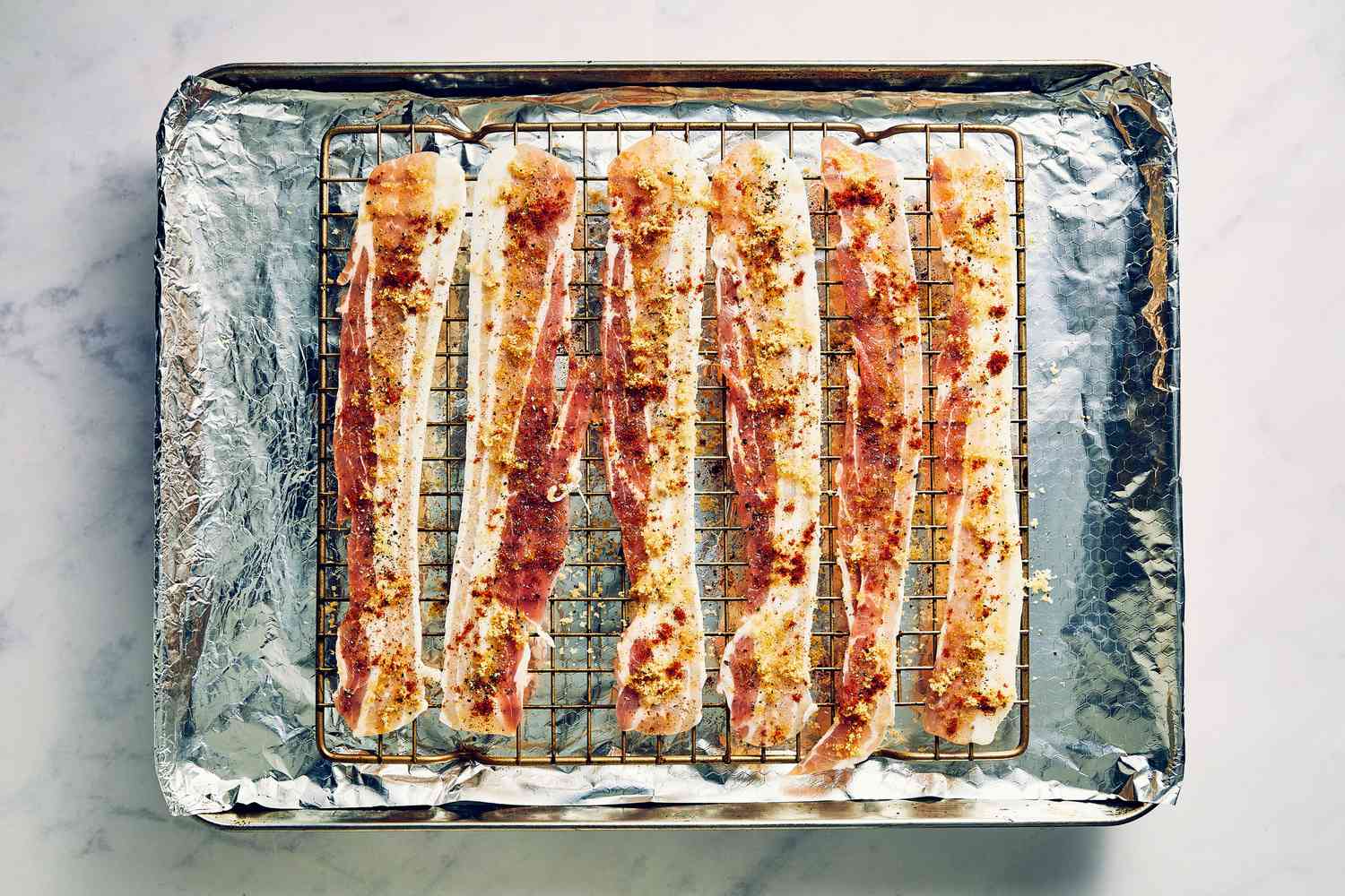 A foil-lined baking sheet with six pieces of bacon topped with brown sugar, black pepper, and cayenne pepper on a baking rack