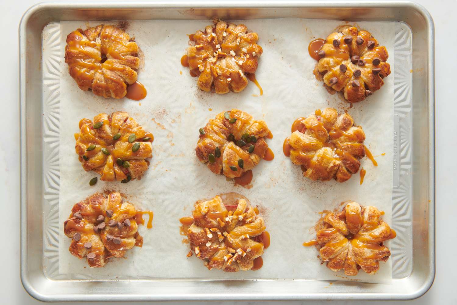 A parchment paper lined baking sheet with apple puff pastry doughnuts topped with miso caramel sauce and garnished with crushed pretzels, pepitas, and chocolate chips
