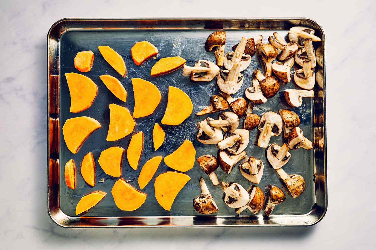 A sheet pan with one side with sliced half moon sweet potato pieces, tossed in oil and seasoned with salt, and the other half with cremini mushrooms tossed in oil and seasoned with salt