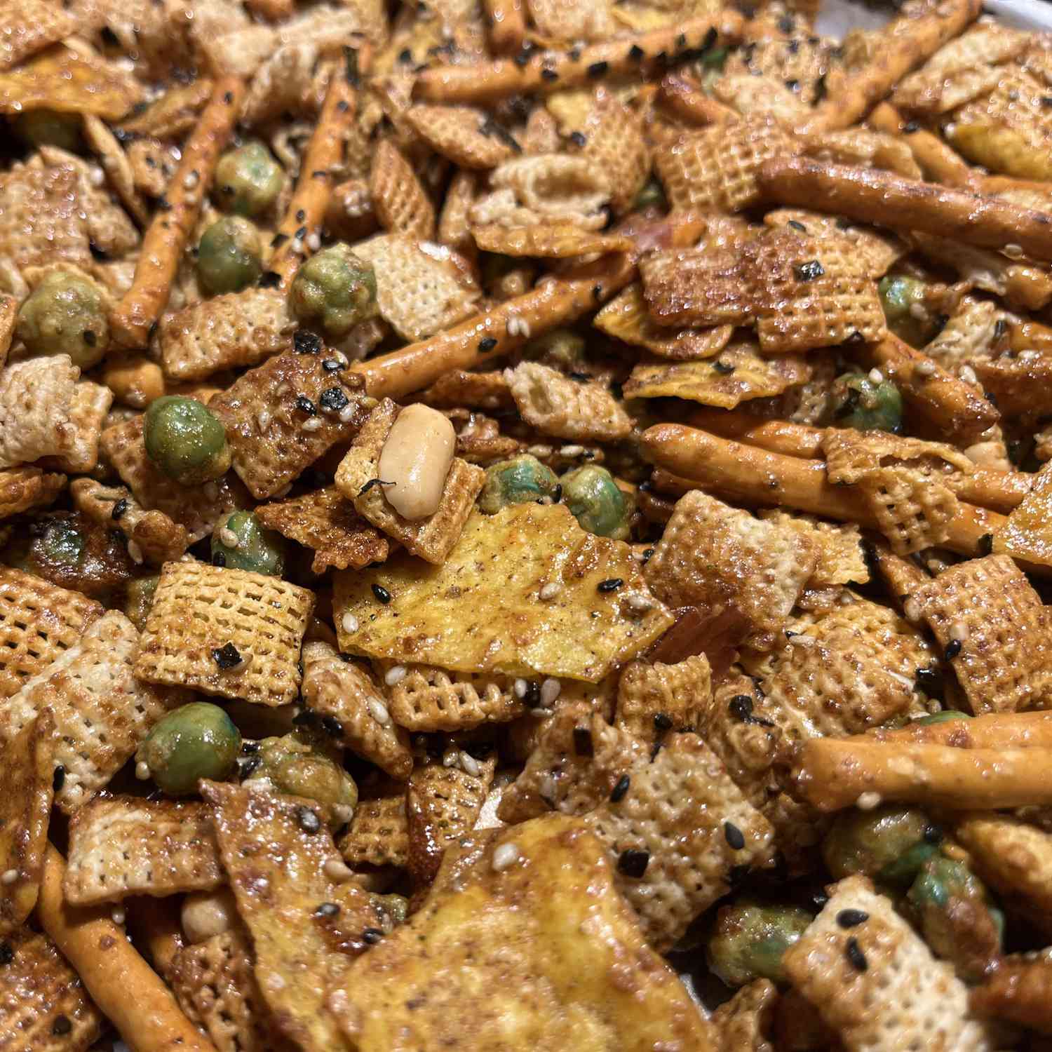 A jumble of golden brow Chex cereal, pretzel rods, nuts, and wasabi peas