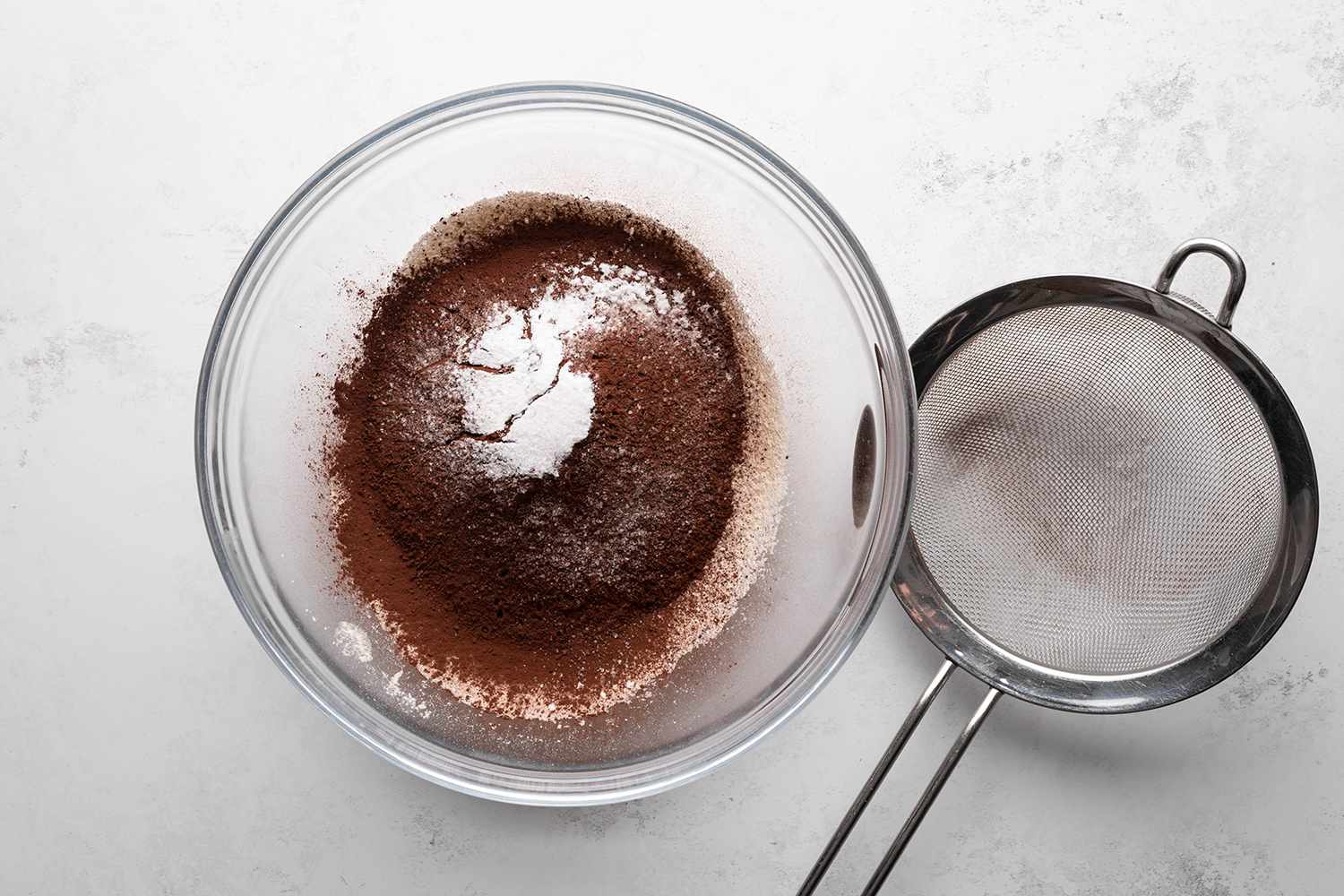Flour, cocoa powder, baking soda, and salt in a glass bowl, next to a mesh strainer