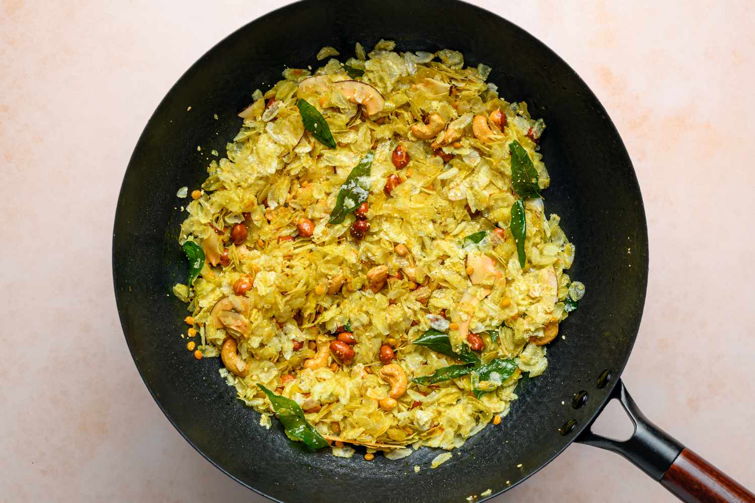 A large wok with a yellowed poha mixture, including the crispy nuts and seeds