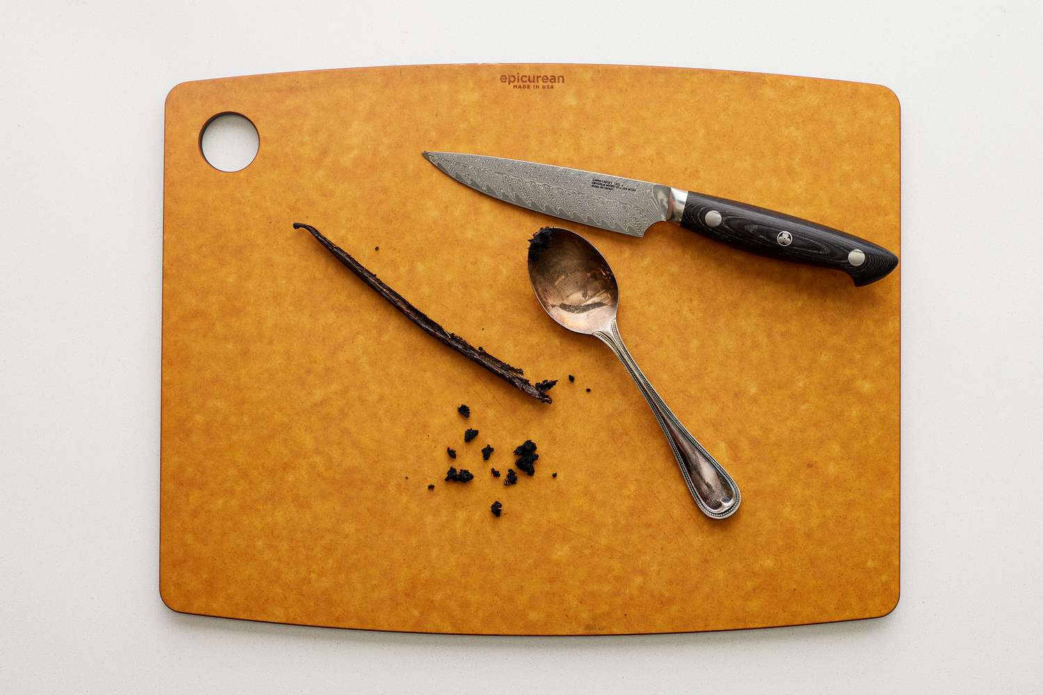 A cutting board with a knife and a spoon, and a vanilla bean pod, cut in half, with vanilla seeds removed. 