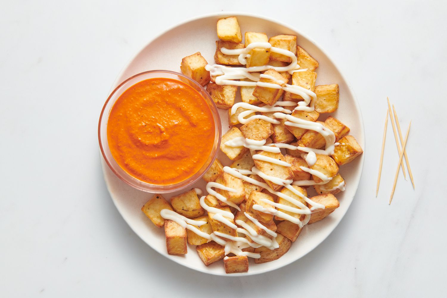 A plater of patatas bravas, drizzled with aioli and served with a small bowl of romesco sauce, with toothpicks on the side. 