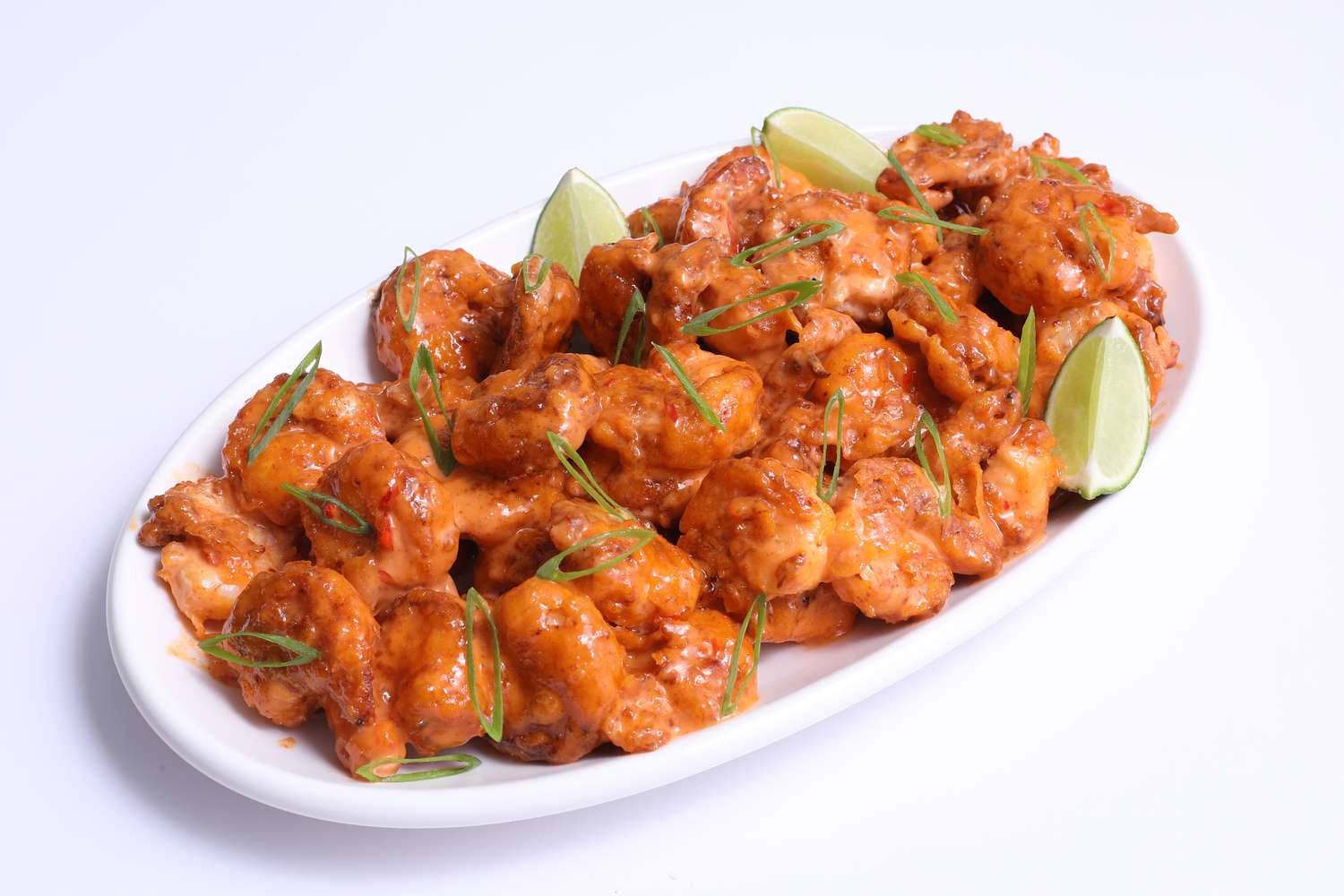 Orange-red colored bang bang shrimp on an oval white platter garnished with sliced scallions and lime wedges