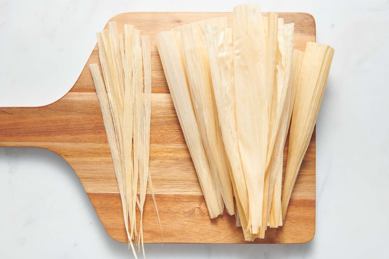 A cutting board with whole corn husks and thin corn husk strips