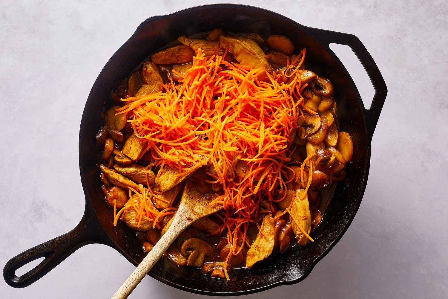 Chicken, shredded carrots, mushrooms and sauce in a cast iron skillet, with a wooden spoon 
