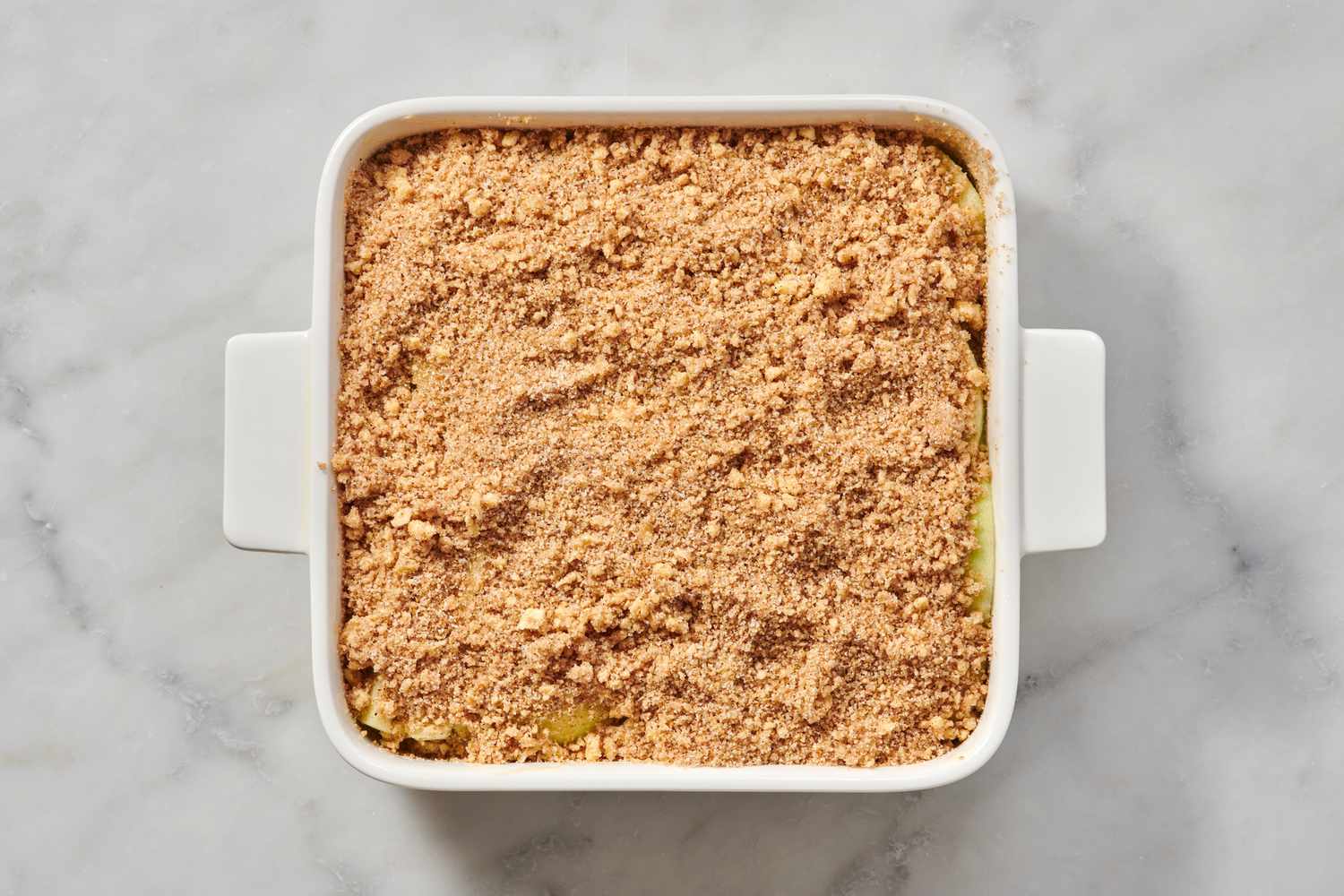 A baking dish lined with sliced apples and seasoned butter-crumb mixture, with a top layer of crumbs