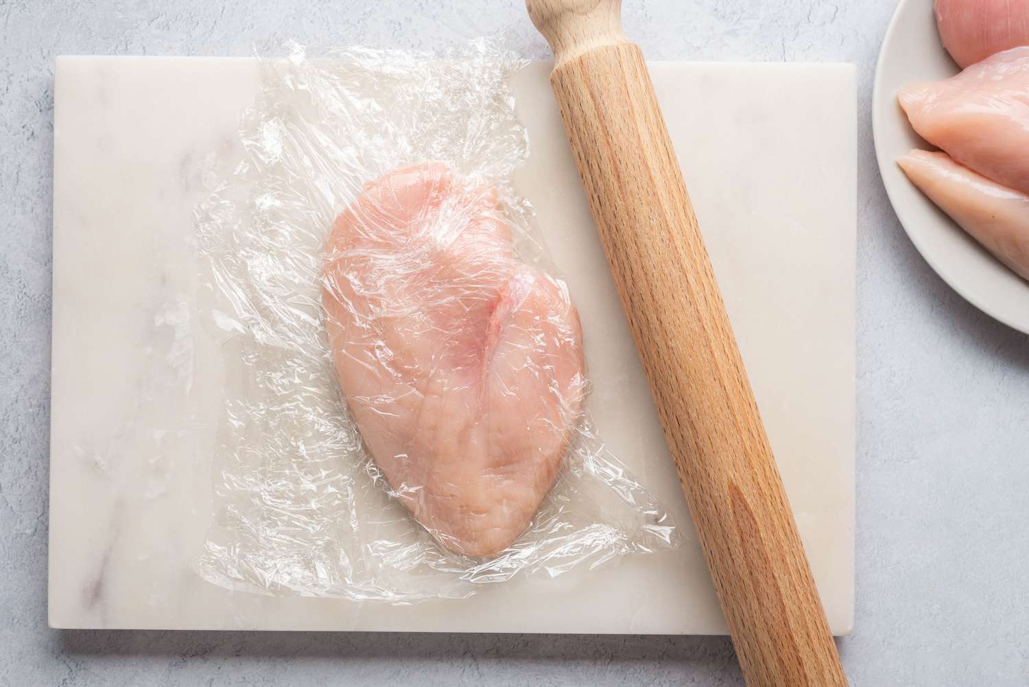 Chicken cutlets pounded thin with a rolling pin on a cutting board with plastic wrap on top