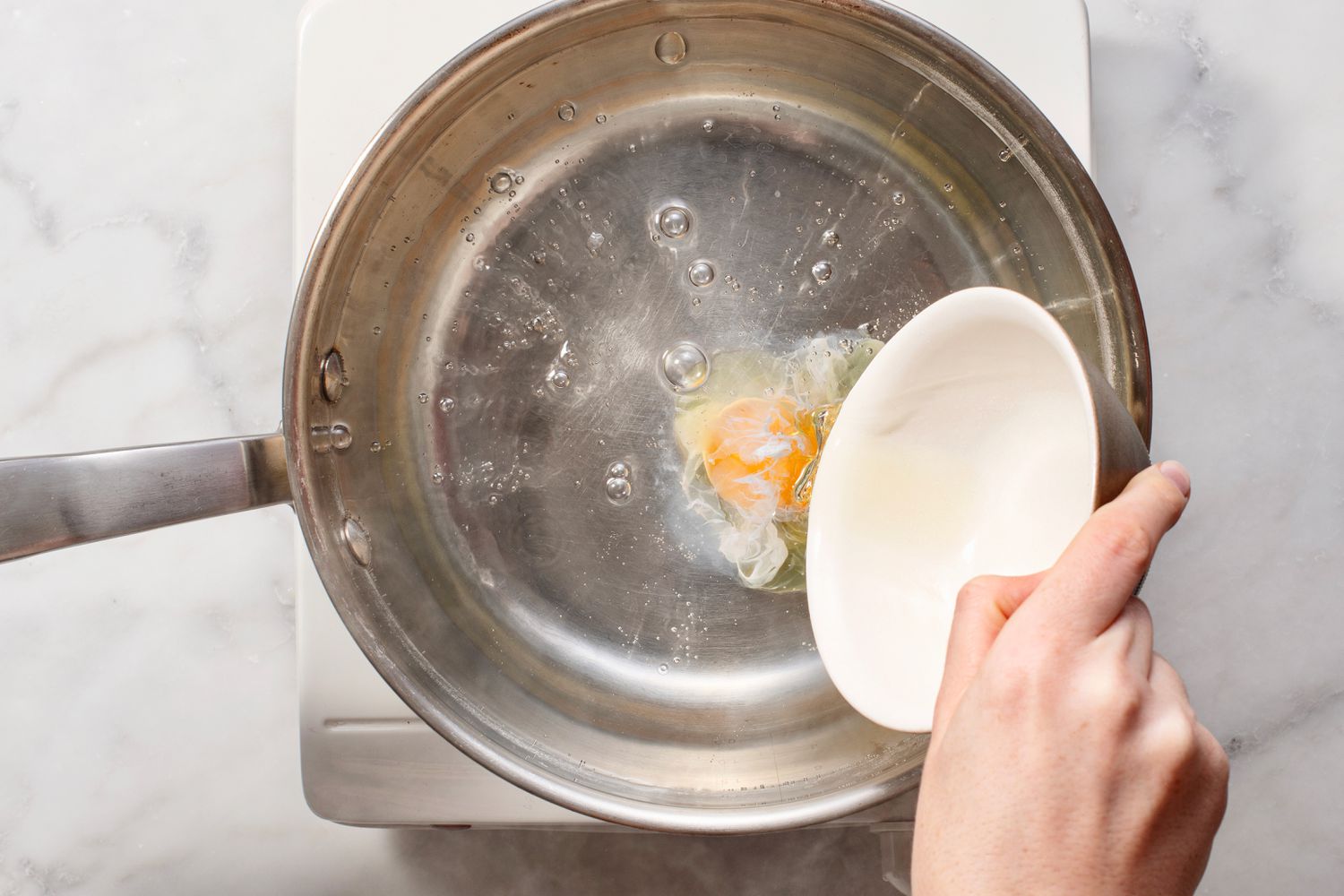 A hand tipping an egg out of a ramekin into the simmering water-vinegar mixture