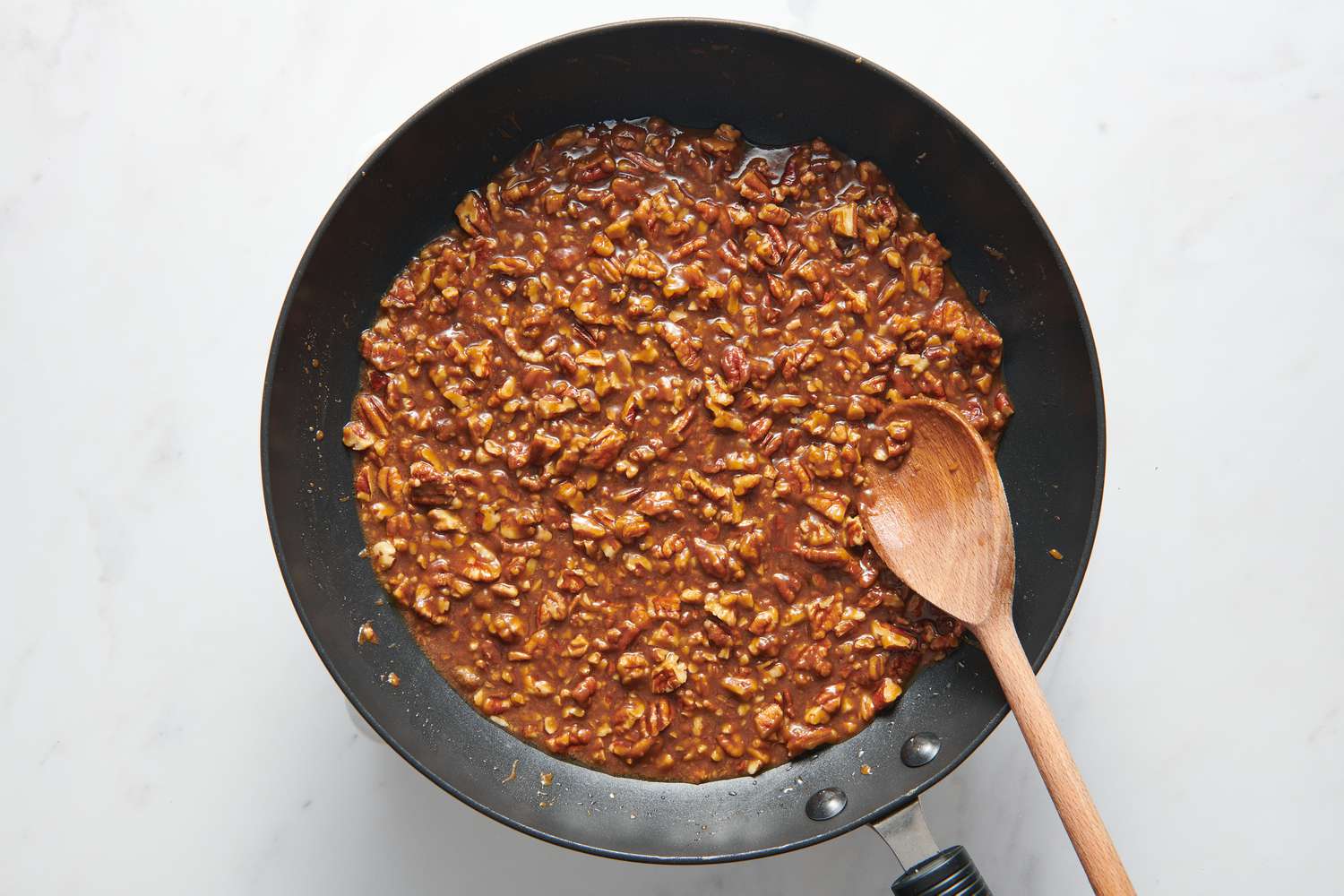 A large pan with chopped pecans, chopped piloncillo, butter, and salt being stirred together with a wooden spoon