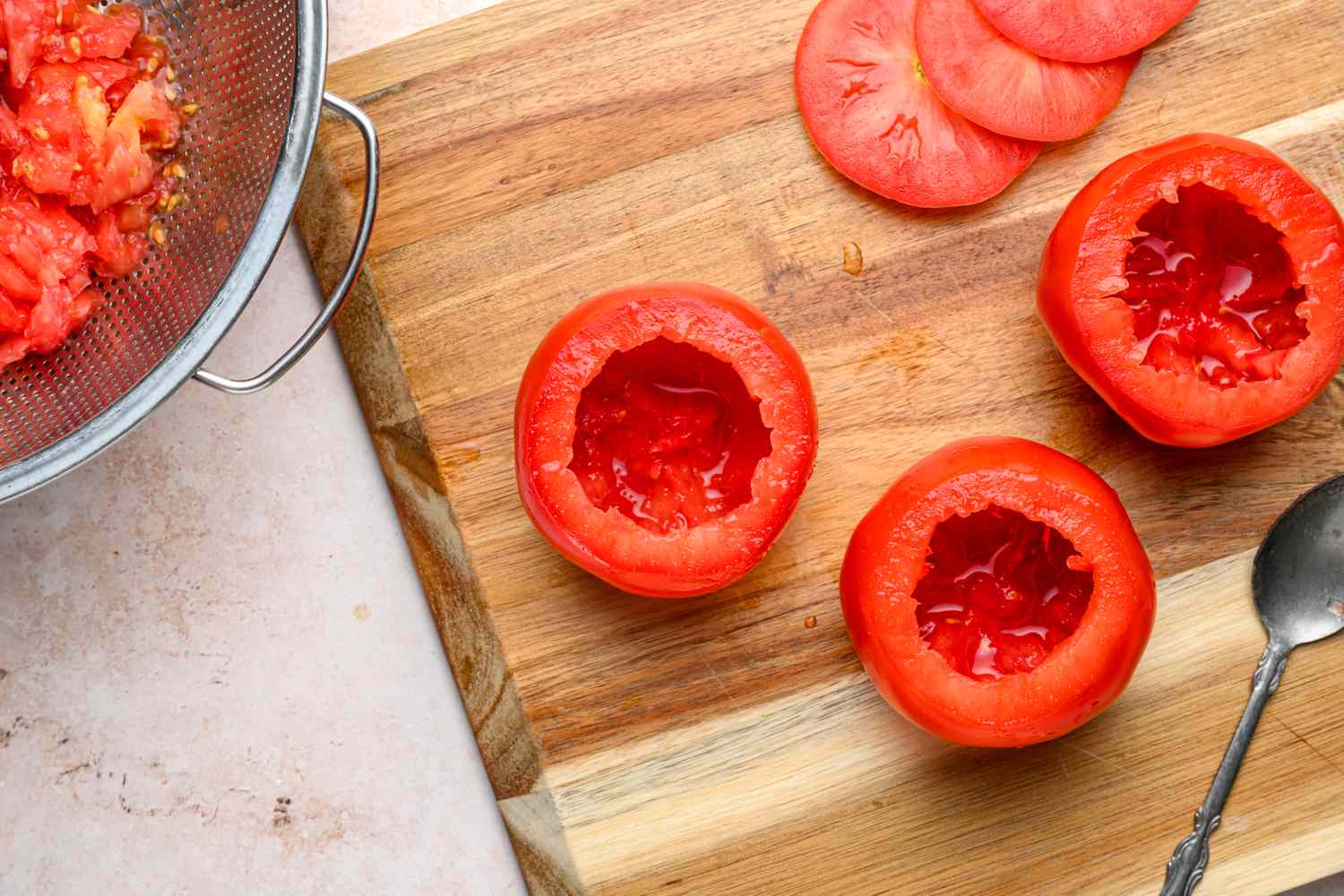 Three tomatoes on a cutting board, hollowed out, with a strainer of tomato seeds