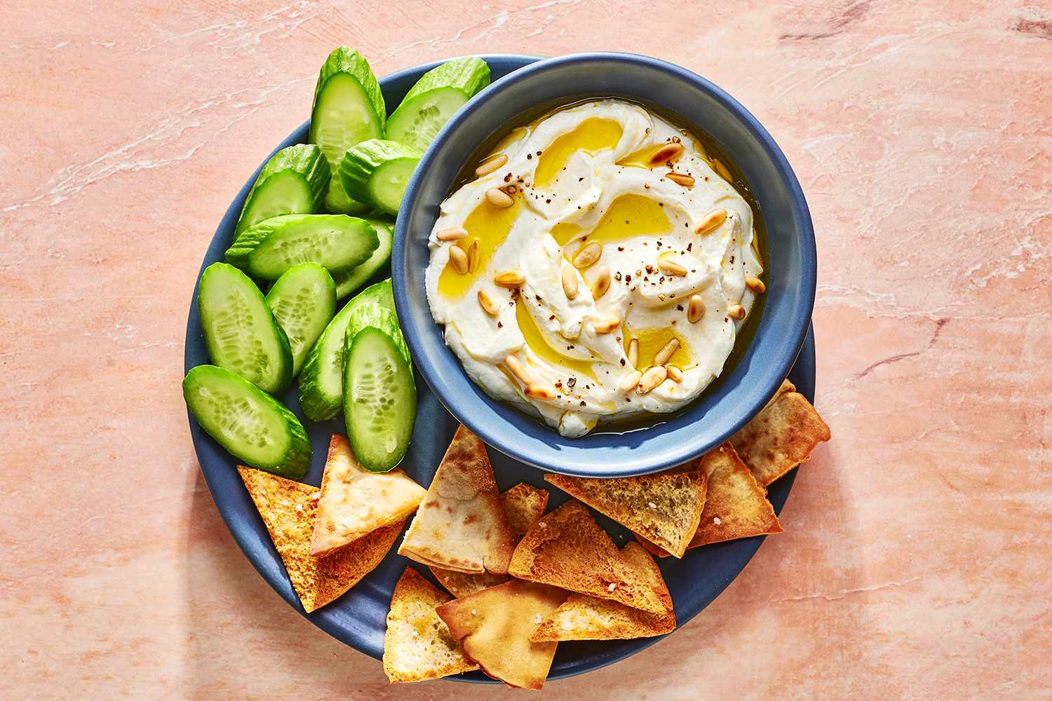A bowl of whipped feta topped with pine nuts, olive oil and black pepper served with cucumbers and pita