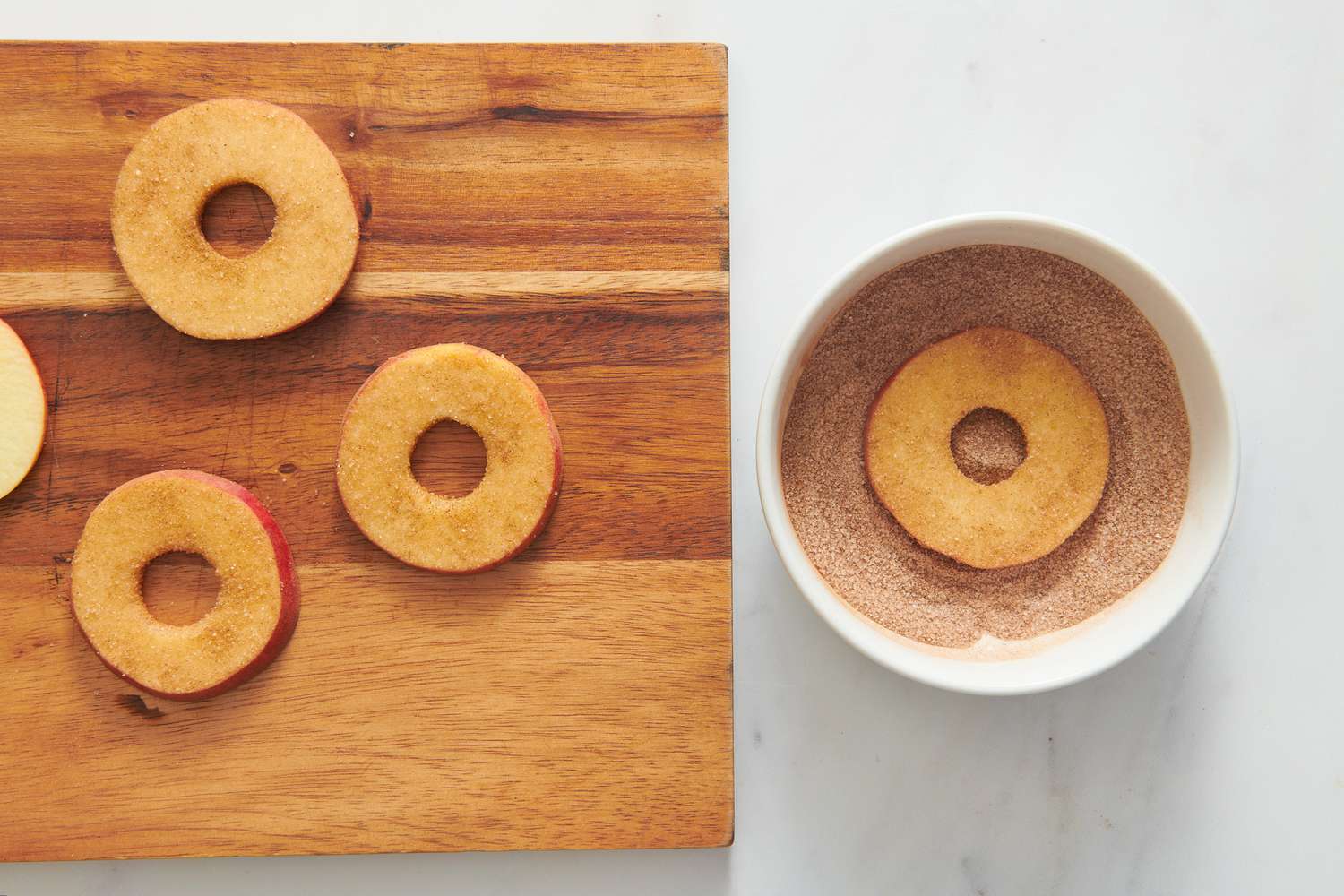 A small bowl of cinnamon sugar with an apple ring being dipped, next to a cutting board with apple rings coated in cinnamon sugar