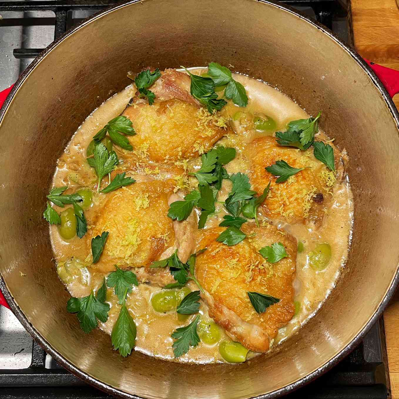 Four browned skin-on chicken thighs in a creamy sauce with green olives; garnished with lemon zest and whole parsley leaves