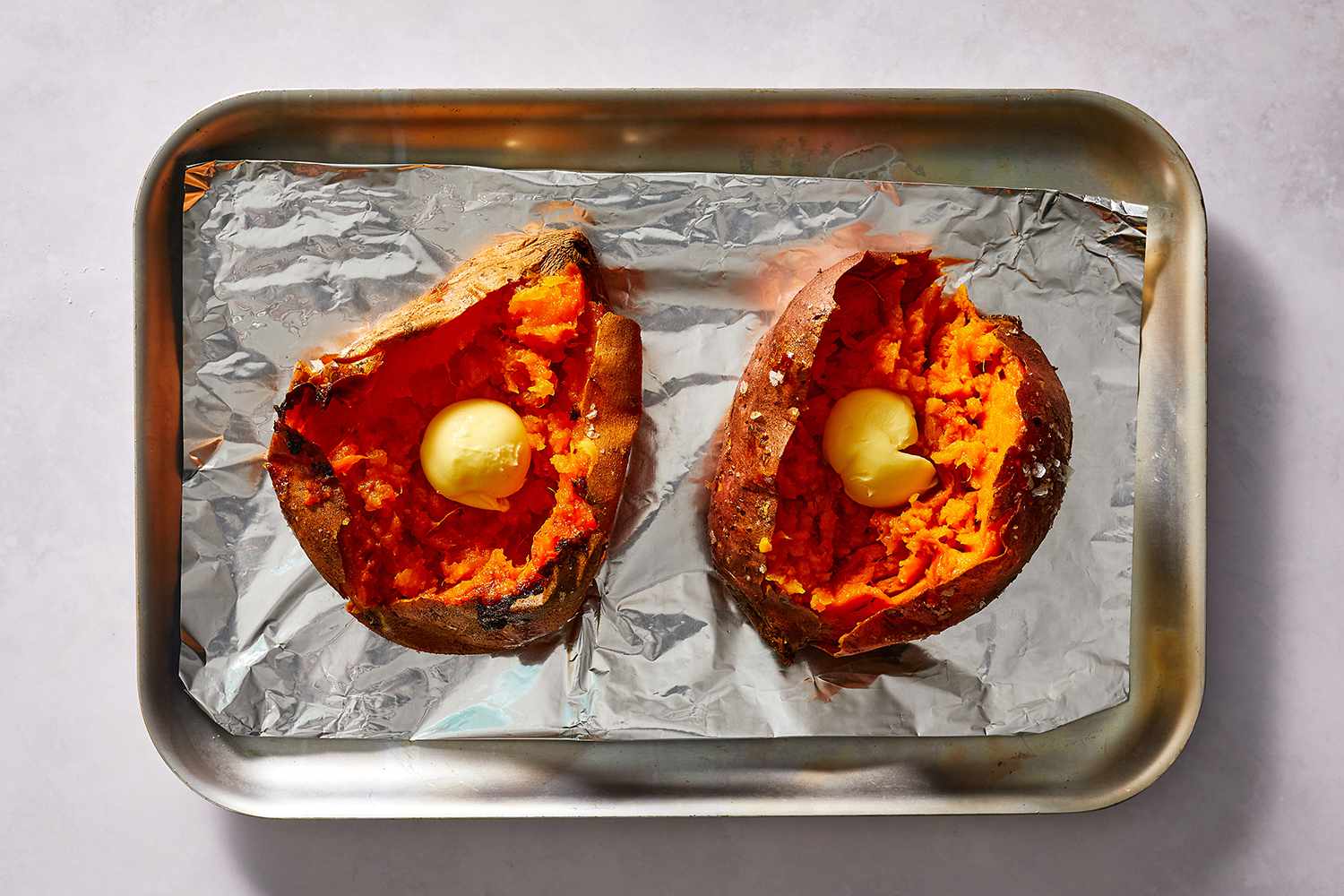 Sweet potatoes topped with butter on an aluminum foil lined baking sheet 