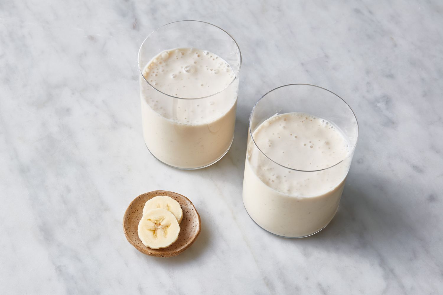 Two glasses of banana smoothie with a small dish with two pieces of sliced banana
