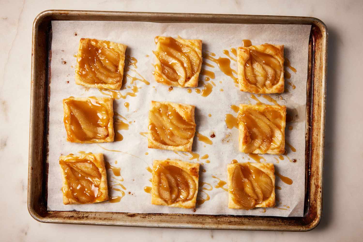A parchment paper-lined baking sheet with nine individual pear tarts topped with brown sugar sauce