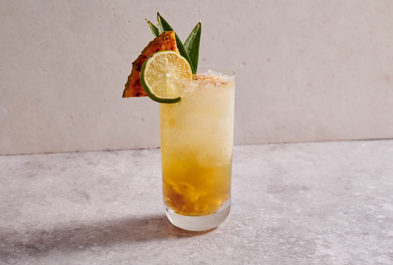 A grilled pineapple chartreuse swizzle cocktail, garnished with cinnamon, grilled pineapple, lime, and pineapple fronds