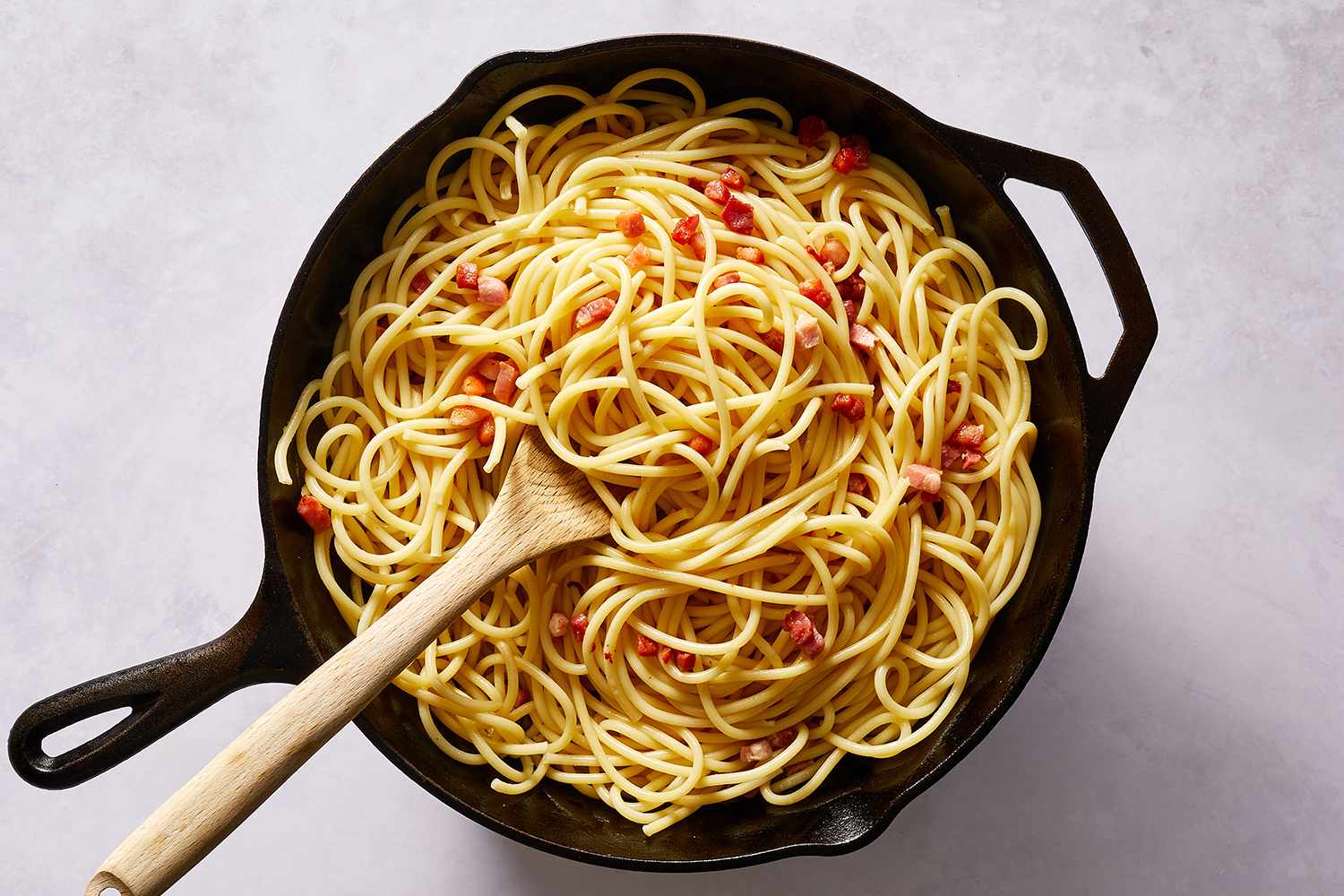 Pancetta and pasta being cooked and stirred together in a cast iron pan