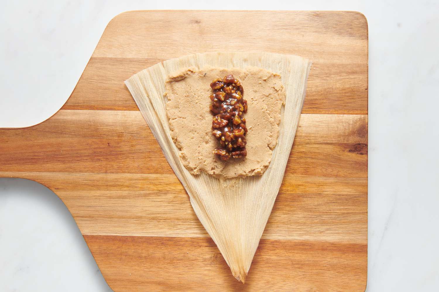 A cutting board with an open corn husk, with a small rectangle of masa harina dough towards the top, topped with a thin portion of pecan pie filling in the middle
