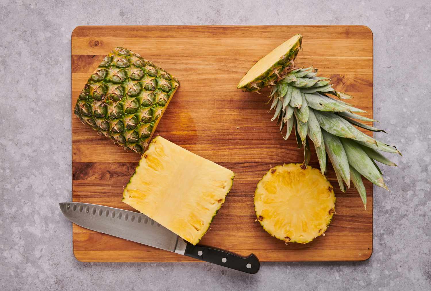 A cutting board with a pineapple cut in half, a bottom slice, and top with fronds
