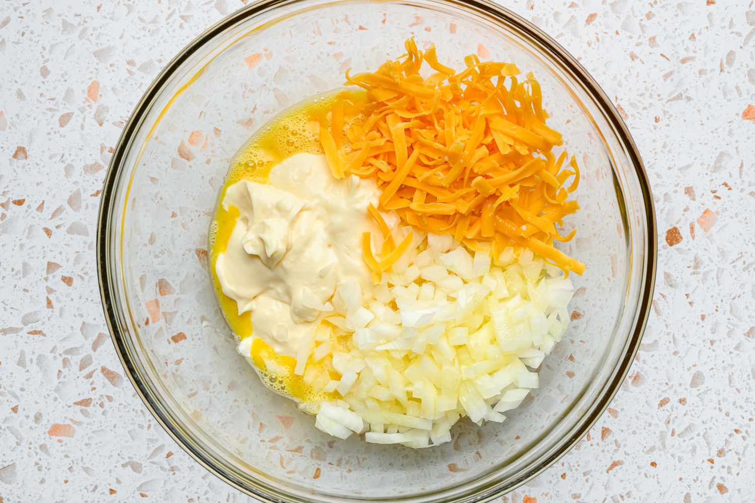 A bowl of mixed egg and sugar, topped with mayonnaise, diced onion, cheddar cheese, and melted butter