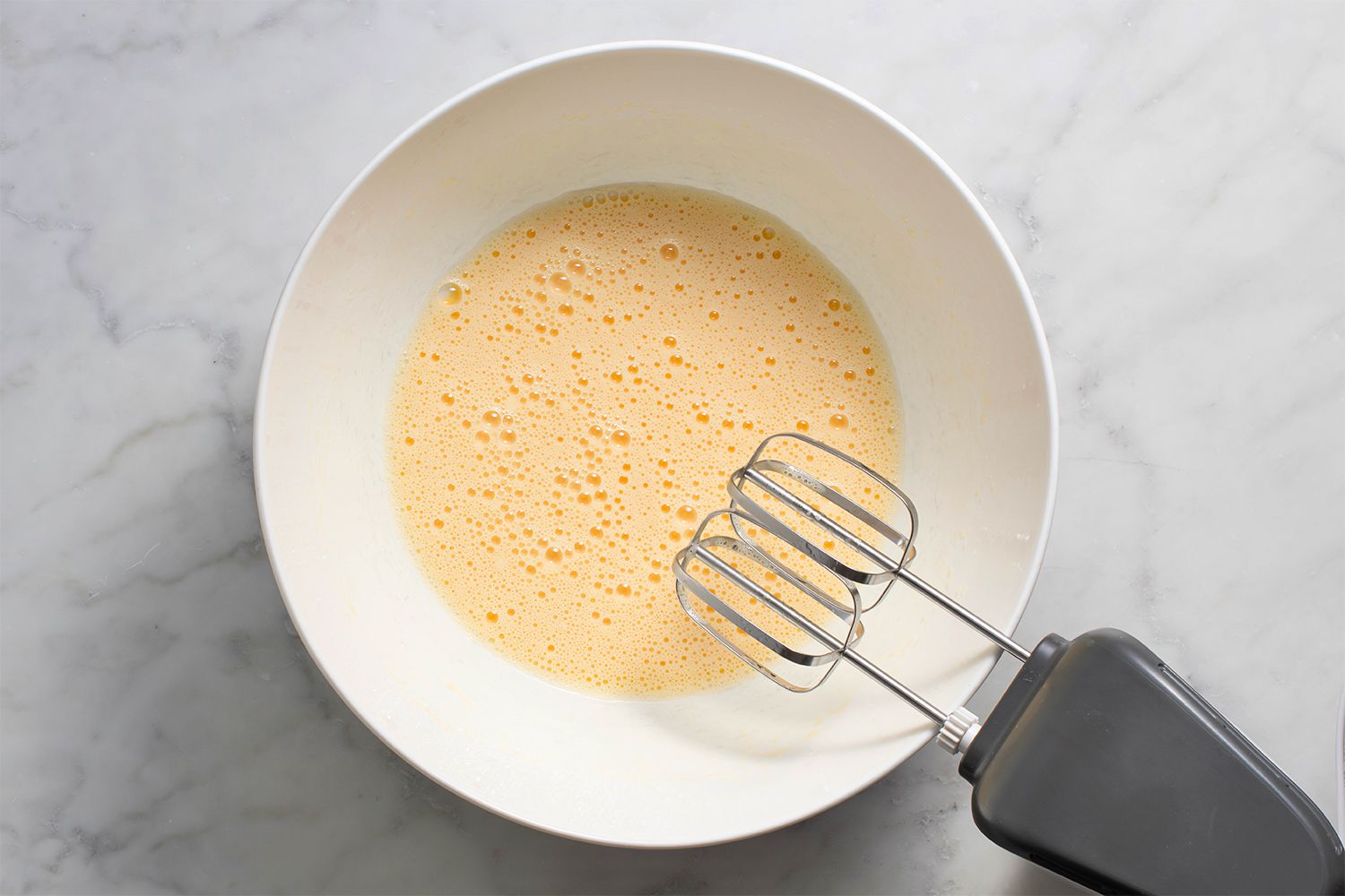 Egg and vegetable oil mixture in a bowl, next to a hand mixer 
