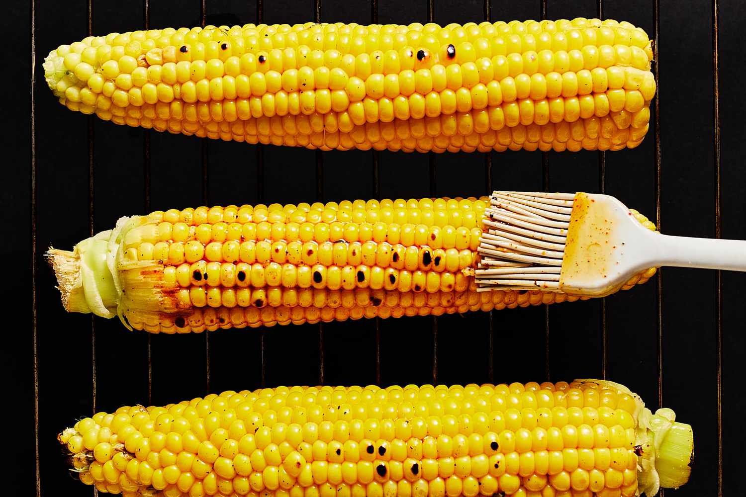 A pastry brush brushing the cajun-oil mixture onto ears of corn on a grill