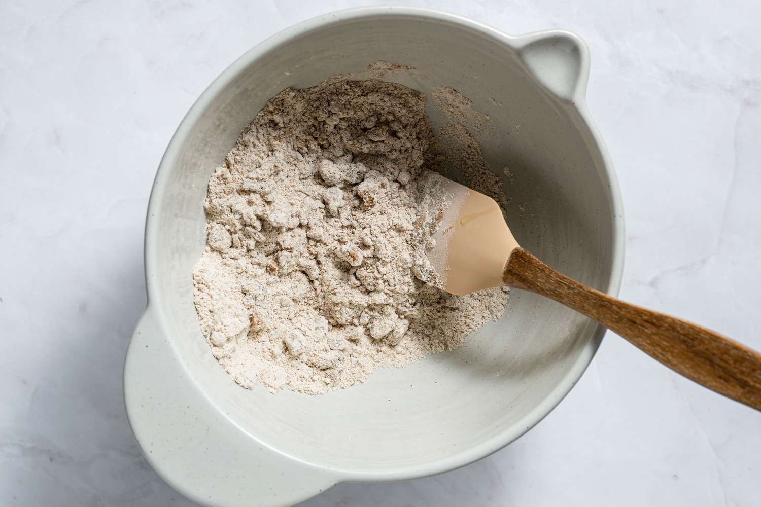 A rubber spatula mixing melted ghee into the bowl of whole wheat flour-carom mixture