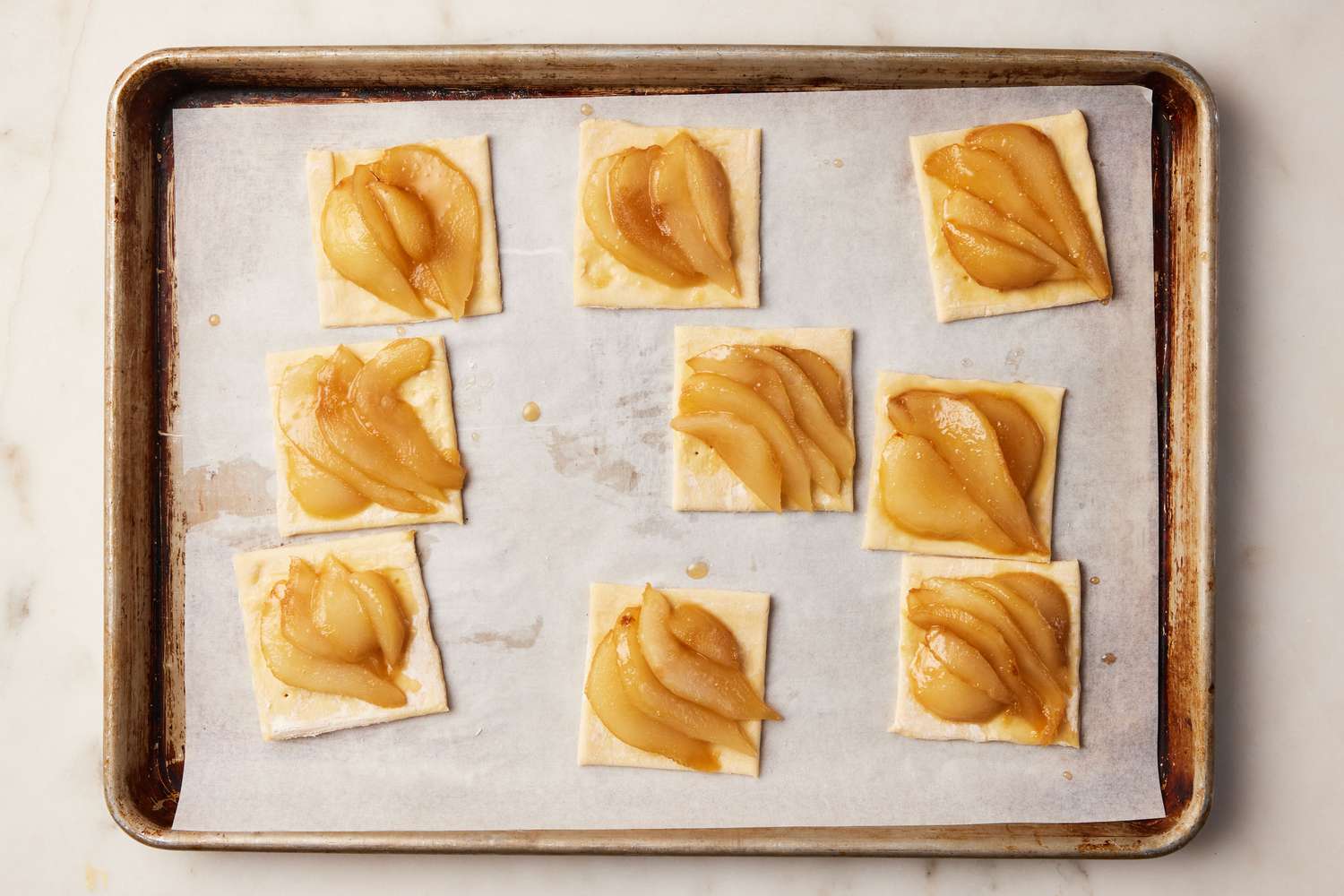 A parchment paper-lined baking sheet with nine squares of puff pastry topped with the cooked pear slices