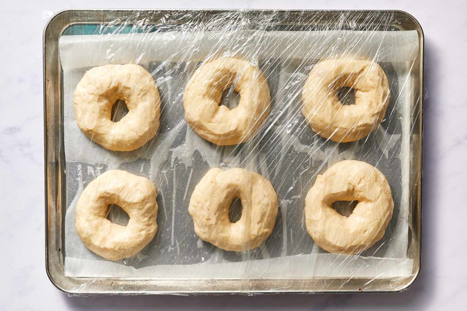 Bagel dough on baking sheet lined with parchment paper and covered with plastic wrap