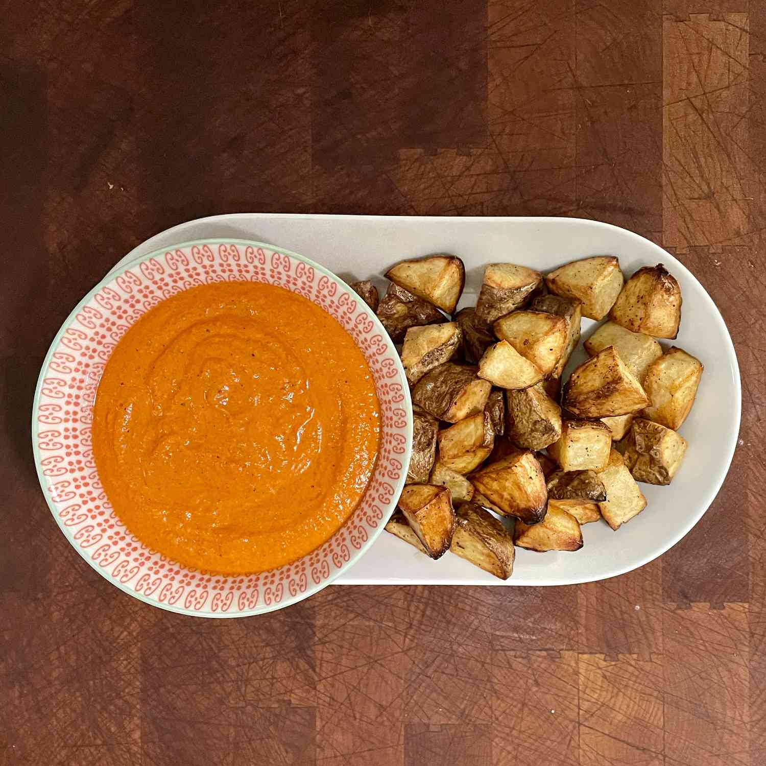 An oblong plate with browned cubed patatas bravas on the right side and a bowl of red romsesco sauce on the left