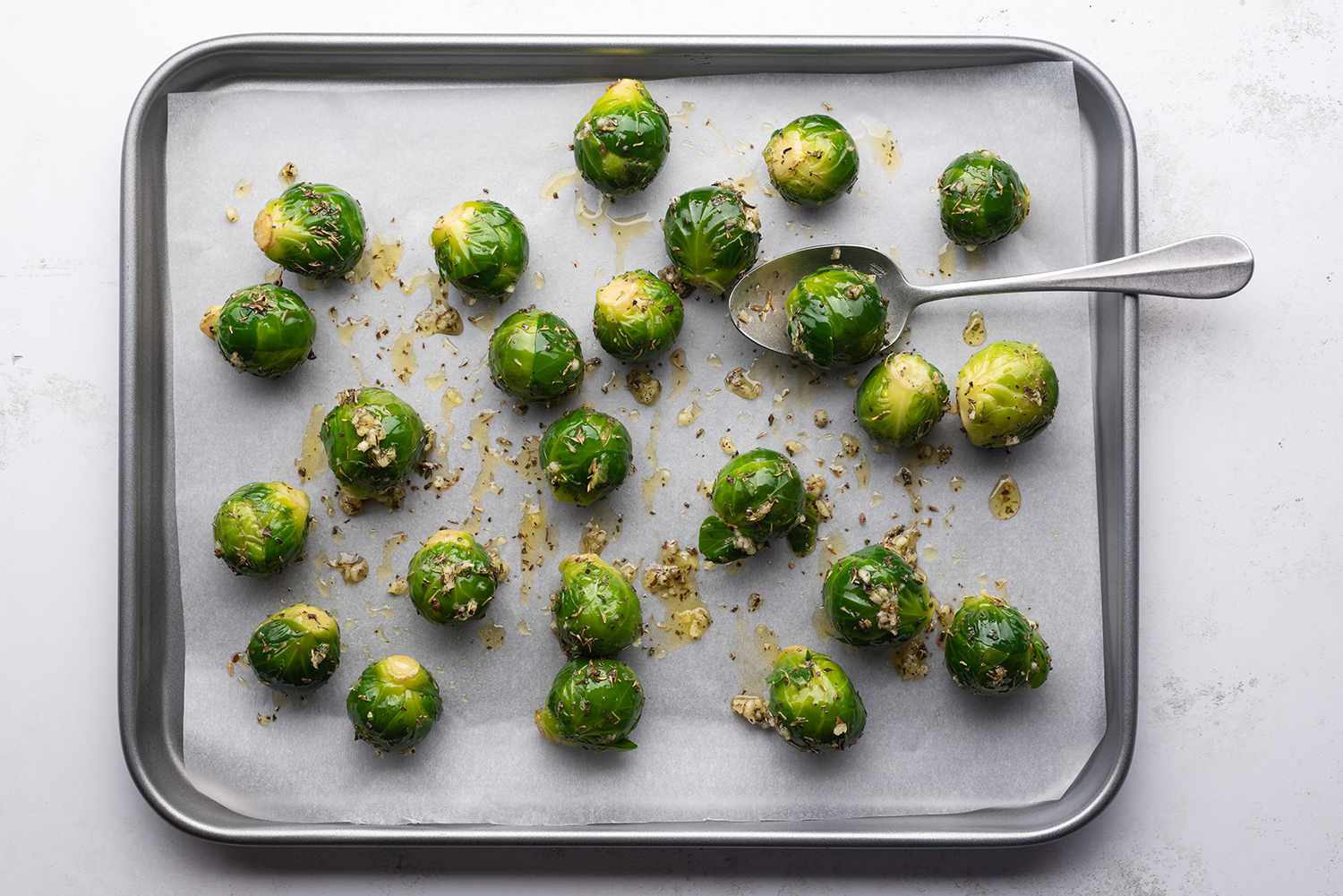 Brussels sprouts with oil, minced garlic, thyme, and basil on a parchment paper lined baking sheet, with a spoon