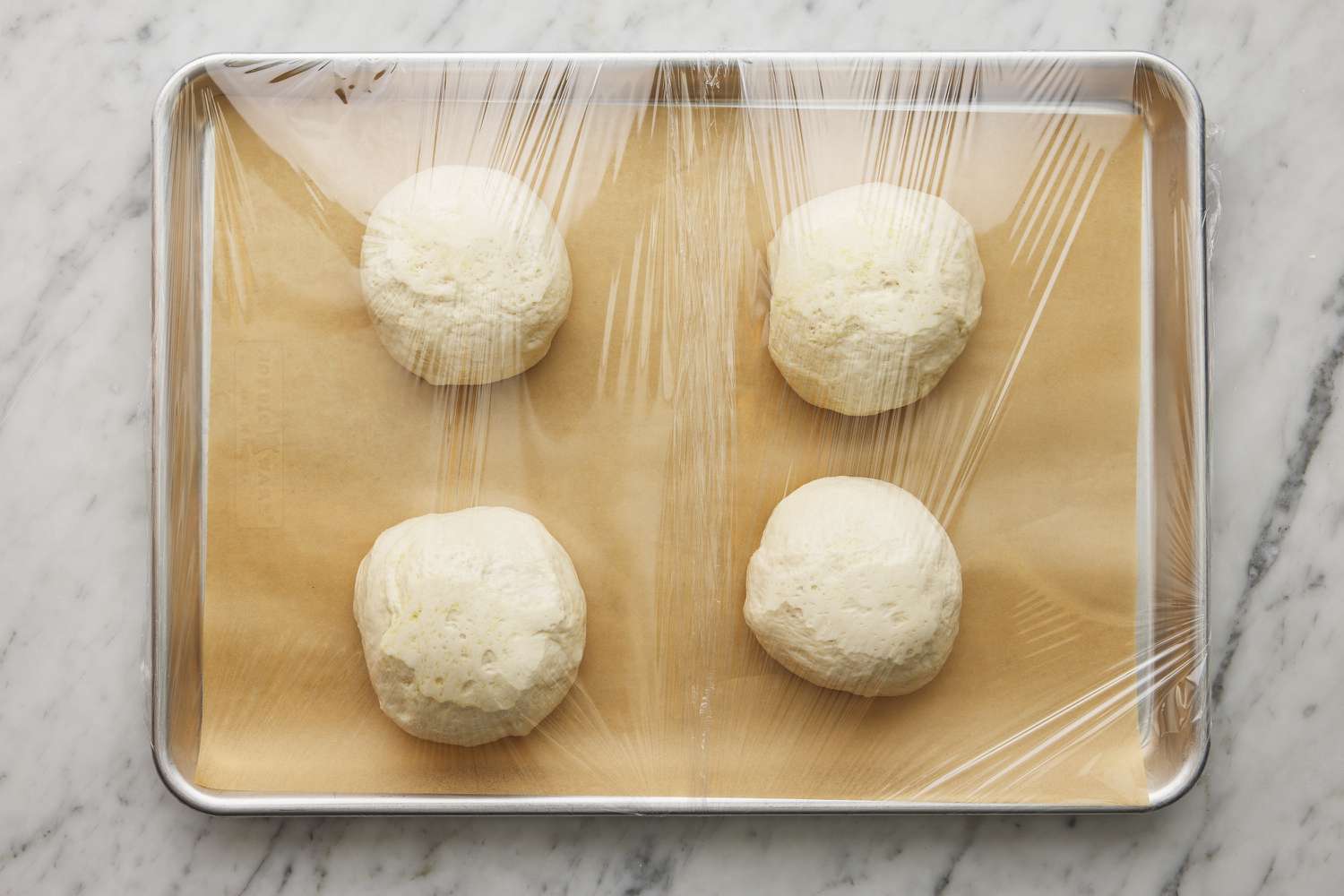 balls of calzone dough resting on parchment lined baking sheet covered with plastic wrap