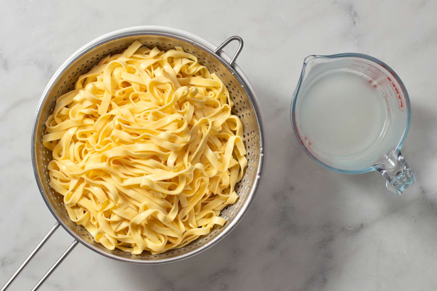 Cooked pasta in a large colander with a measuring cup of pasta water