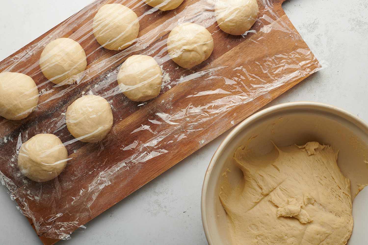 Dough in a bowl, and dough balls on a wooden cutting board covered with plastic wrap 