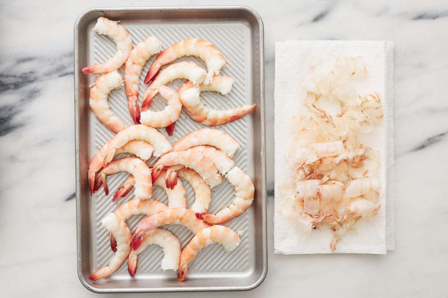 A sheet pan of peeled, jumbo shrimp next to a paper towel with shrimp shells
