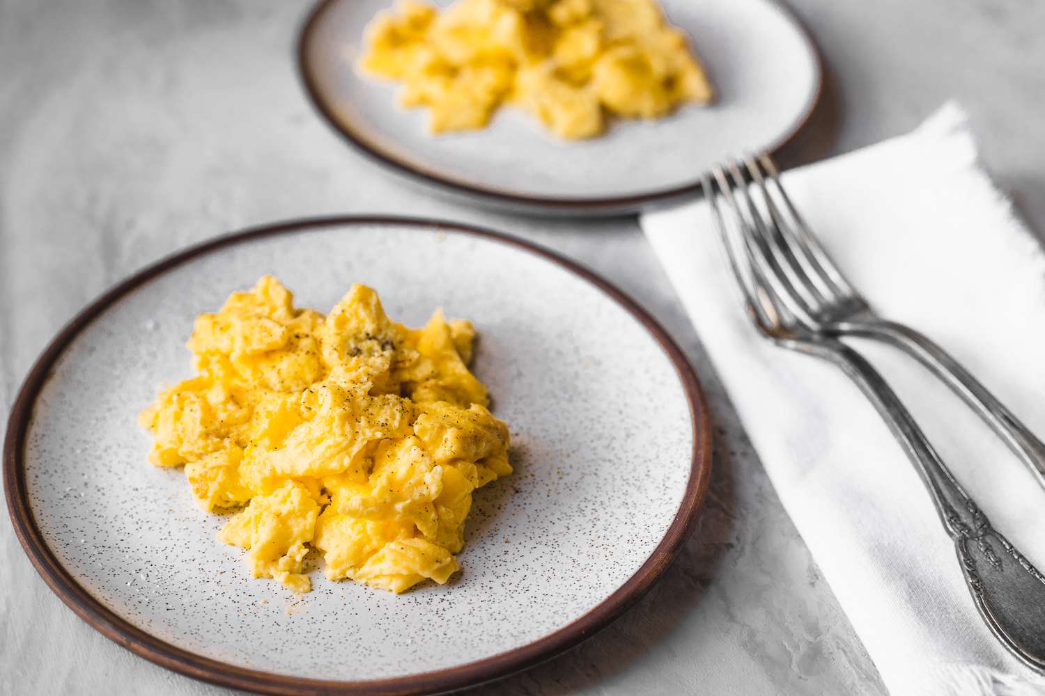 Two plates of fluffy scrambled eggs with two forks and a napkin