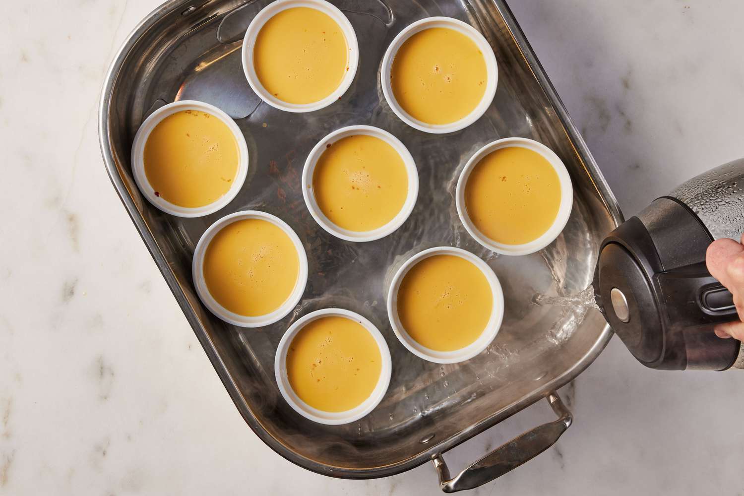 A roasting pan with eight ramekins of raw mango flan being filled with hot water from an electric kettle