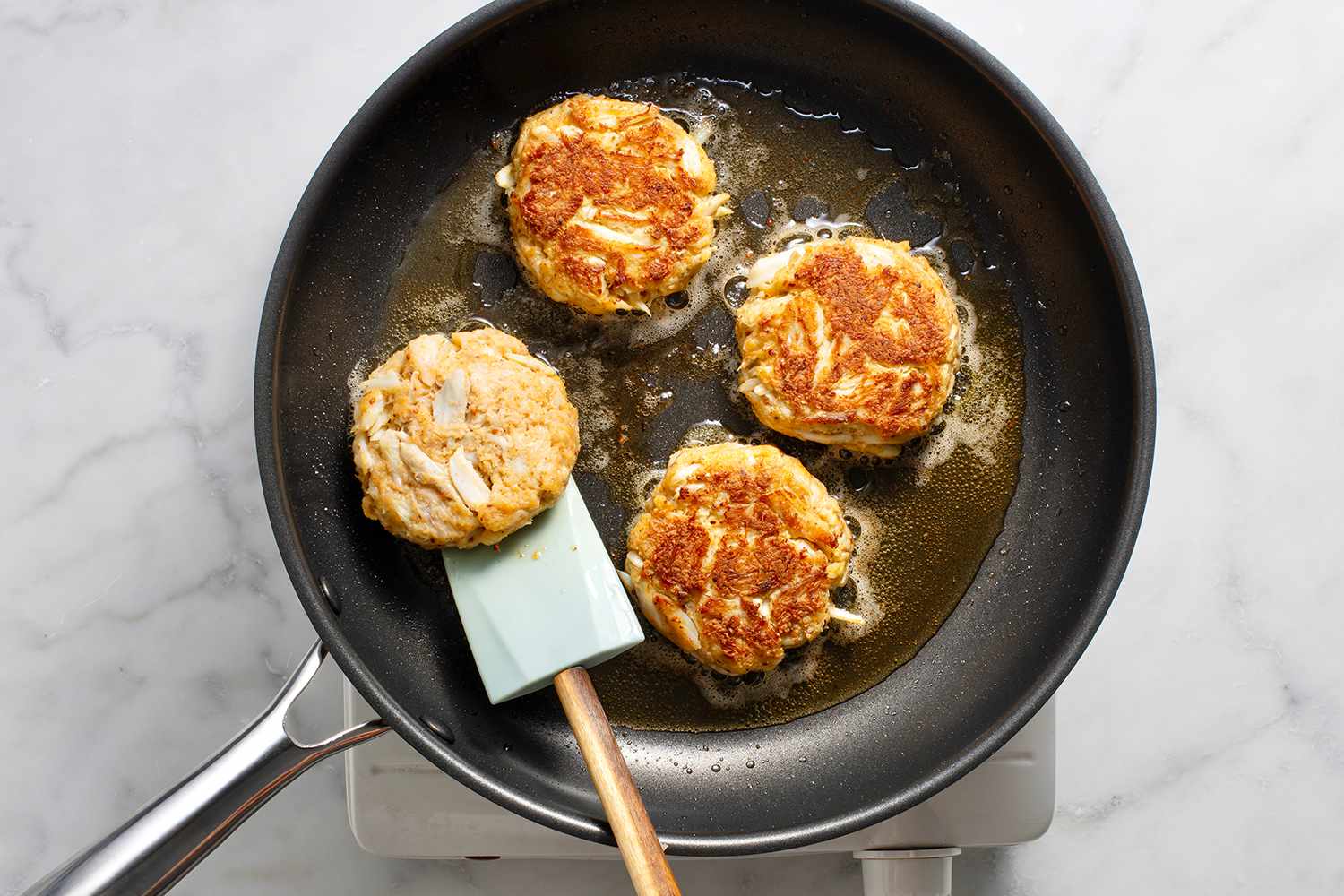 Crab cakes cooking in a pan on a burner, with a spatula 