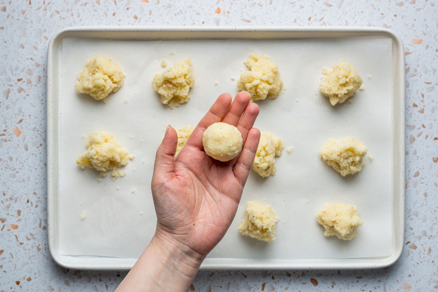 A hand holding a perfectly round portion of the coconut laddoo mixture, over a parchment paper-lined baking sheet of portioned dough