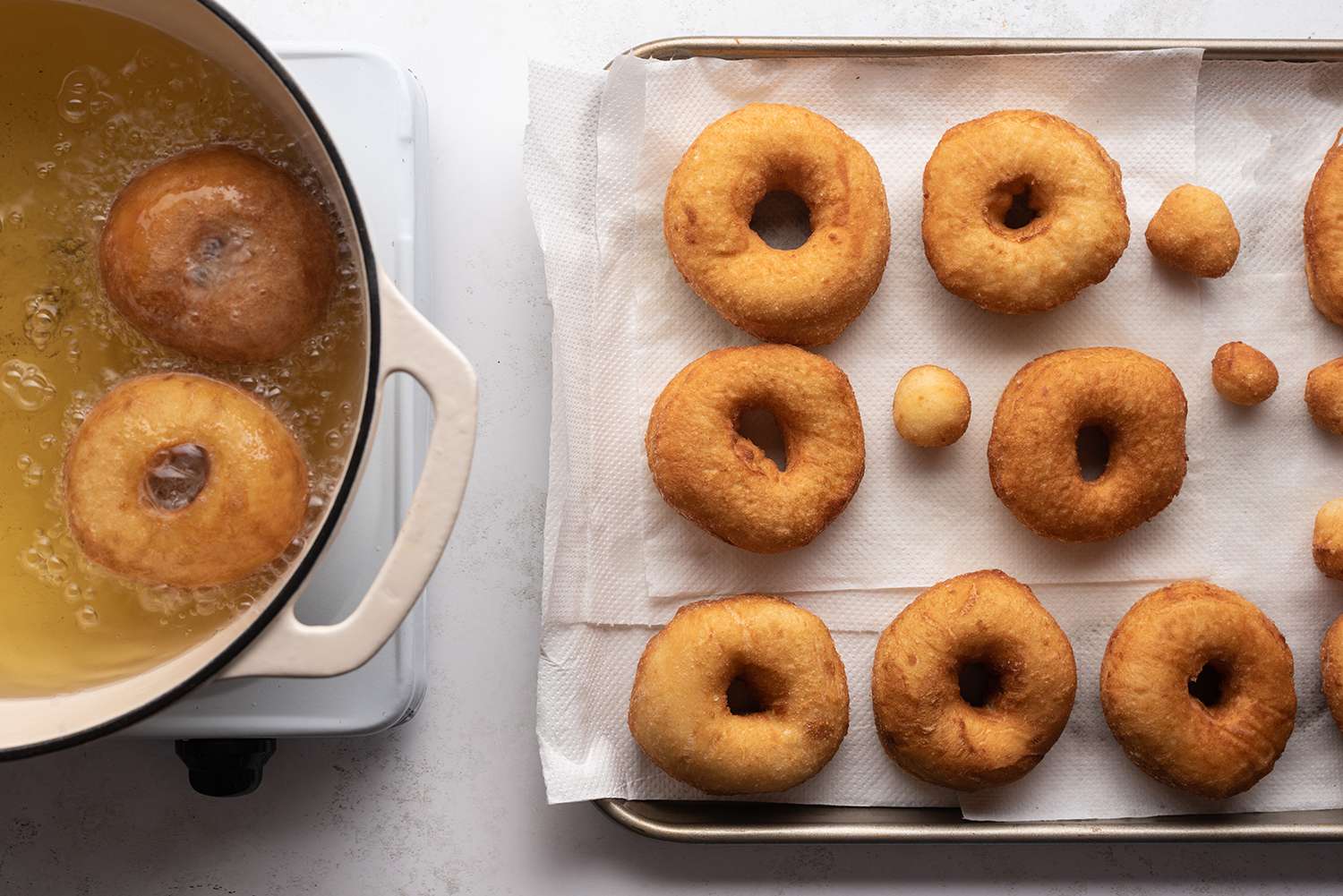 Doughnuts frying in a pot with oil, and doughnuts on a lined baking sheet 