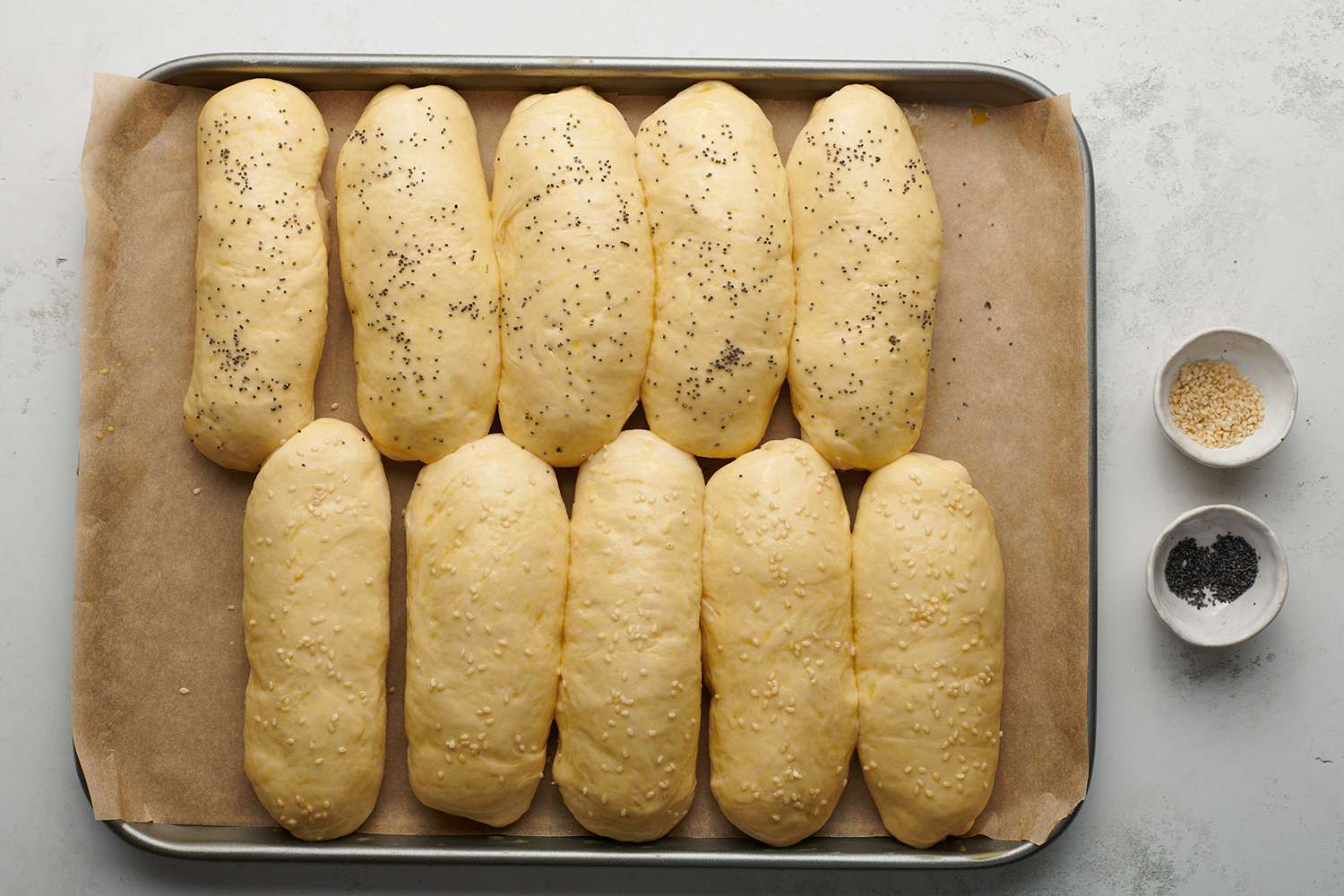 Hot dog bun dough on a parchment paper lined baking sheet, sprinkled sesame seeds and poppy seeds