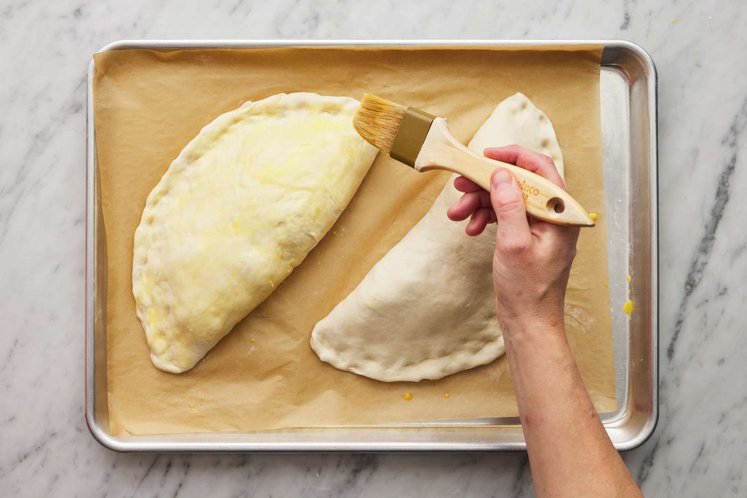 brushing egg wash with a pastry brush on two filled and folded calzones before baking
