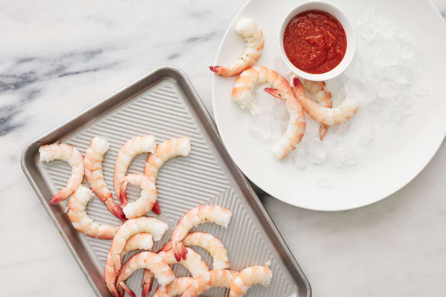 A large serving plate with ice, jumbo shrimp, and a small bowl of cocktail sauce next to a sheet pan of peeled jumbo shrimp