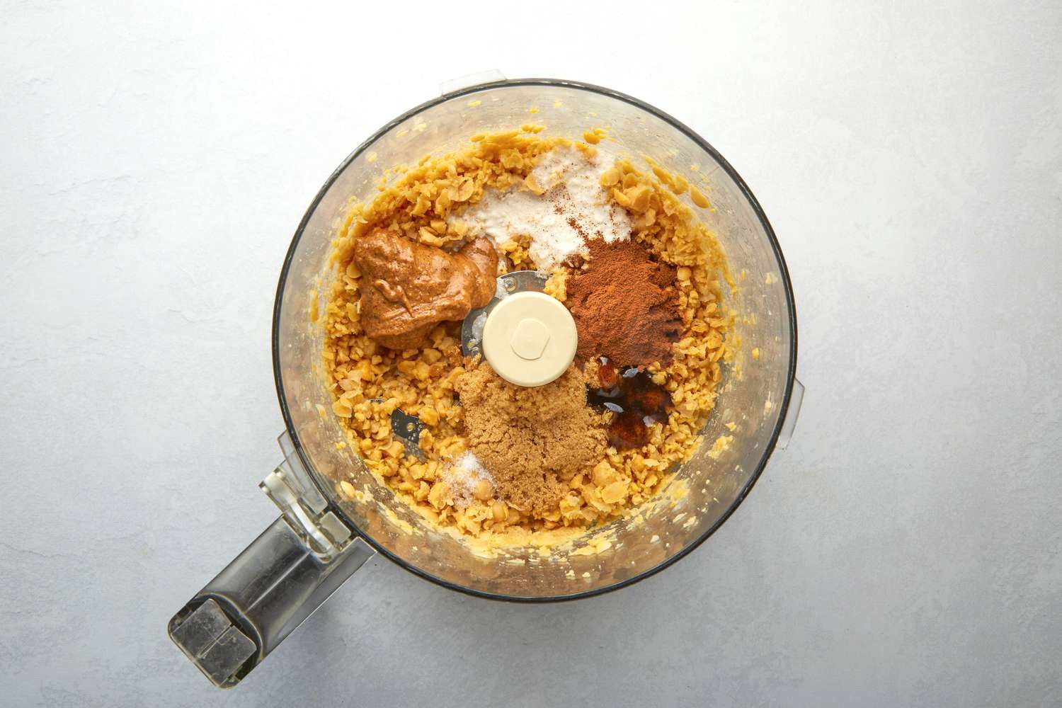 A food processor with crumbled up chickpeas, almond butter, coconut milk, cinnamon, maple syrup, brown sugar, vanilla paste, and salt